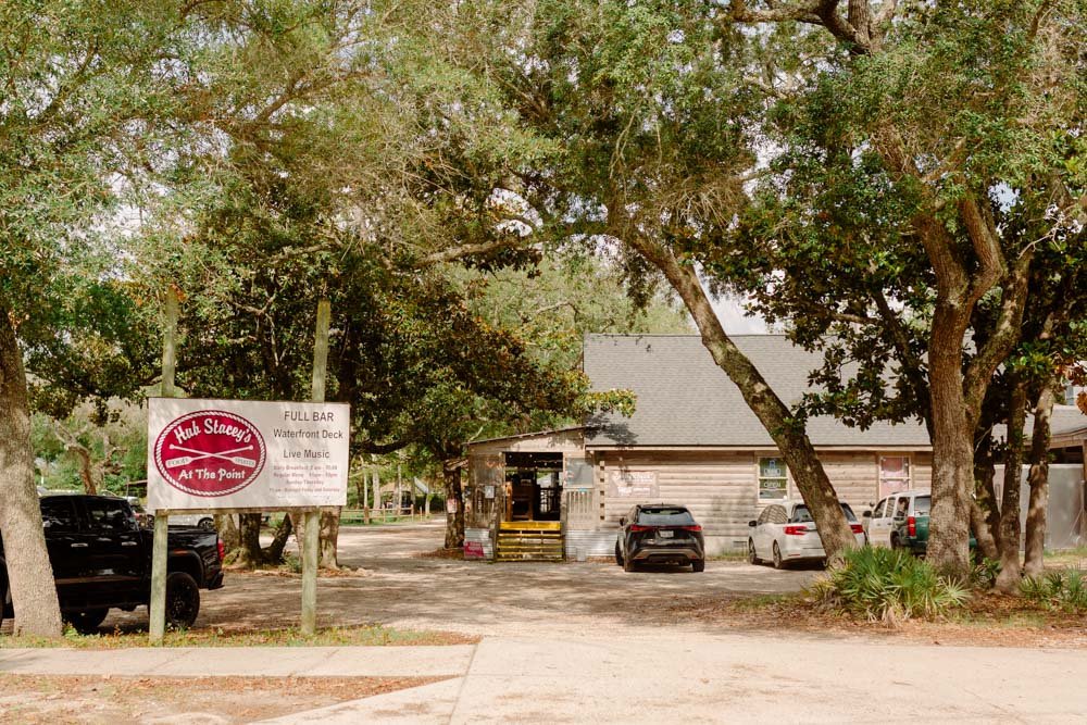 A small wooden restaurant called Hub Stacey's with a sign indicating it's a full bar, waterfront deck, and live music, surrounded by trees and parked cars.