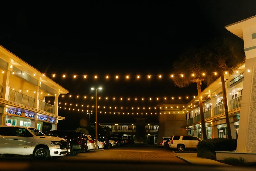 Nighttime scene of outdoor shopping area with string lights overhead, parked cars, palm trees, and multi-story buildings with balconies.