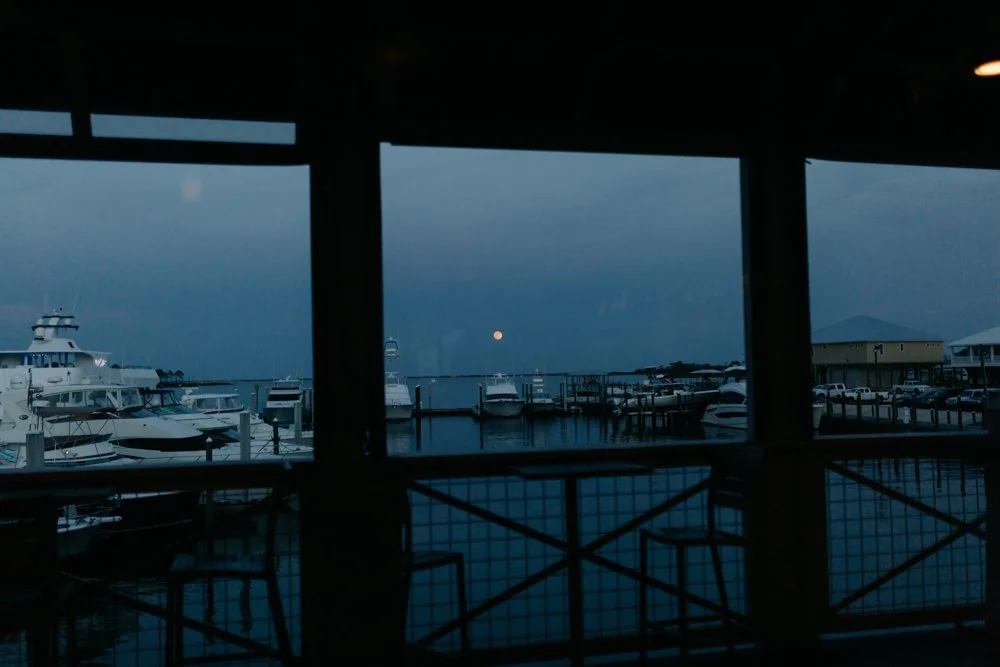 View from a covered marina showing boats docked at the pier during dusk, with the moon rising in the sky.