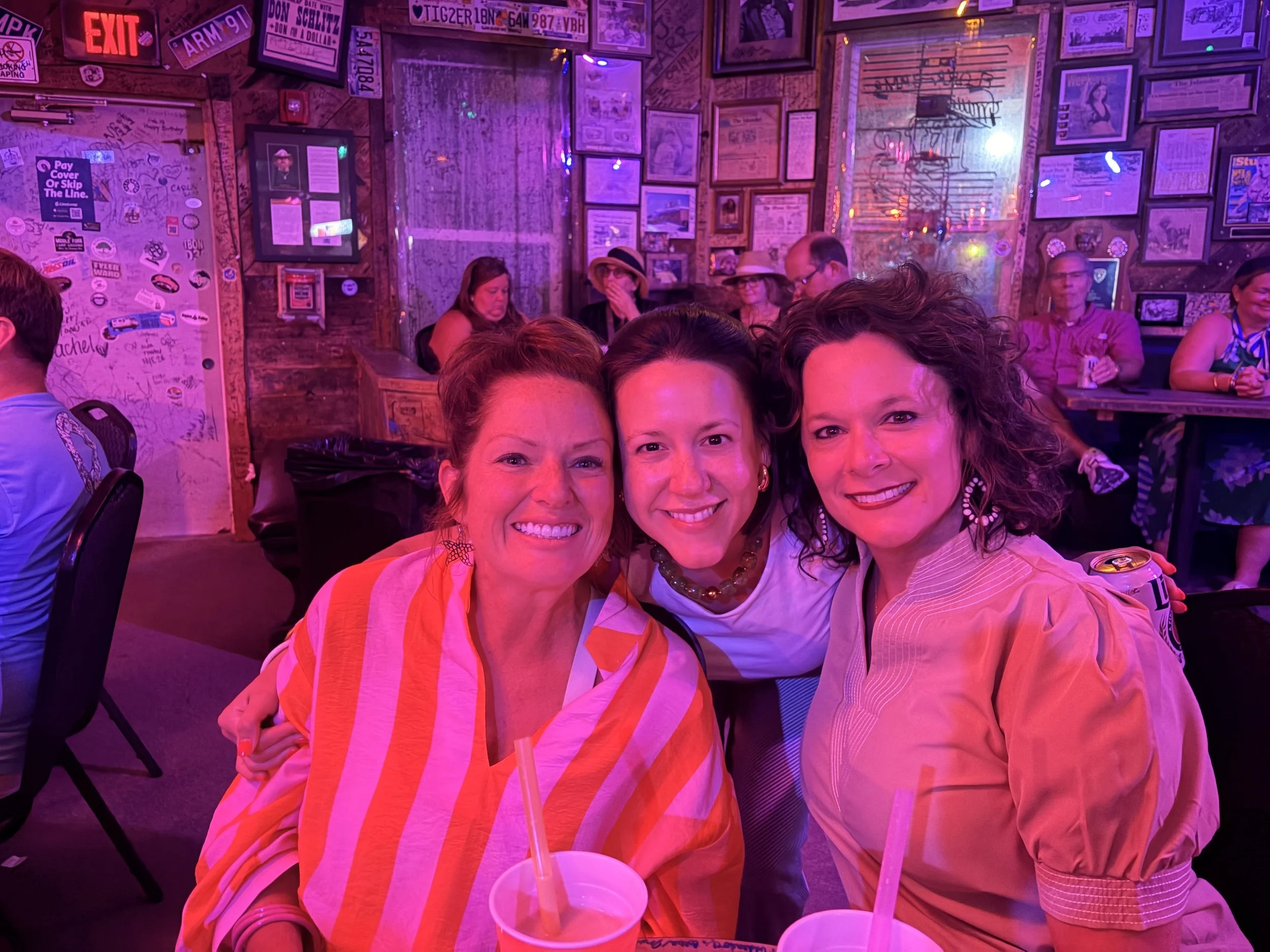 Four women are smiling and posing for a photo in a dimly lit, colorful bar or restaurant. The background features framed pictures, posters, and graffiti on the walls, with purple and pink neon lighting.