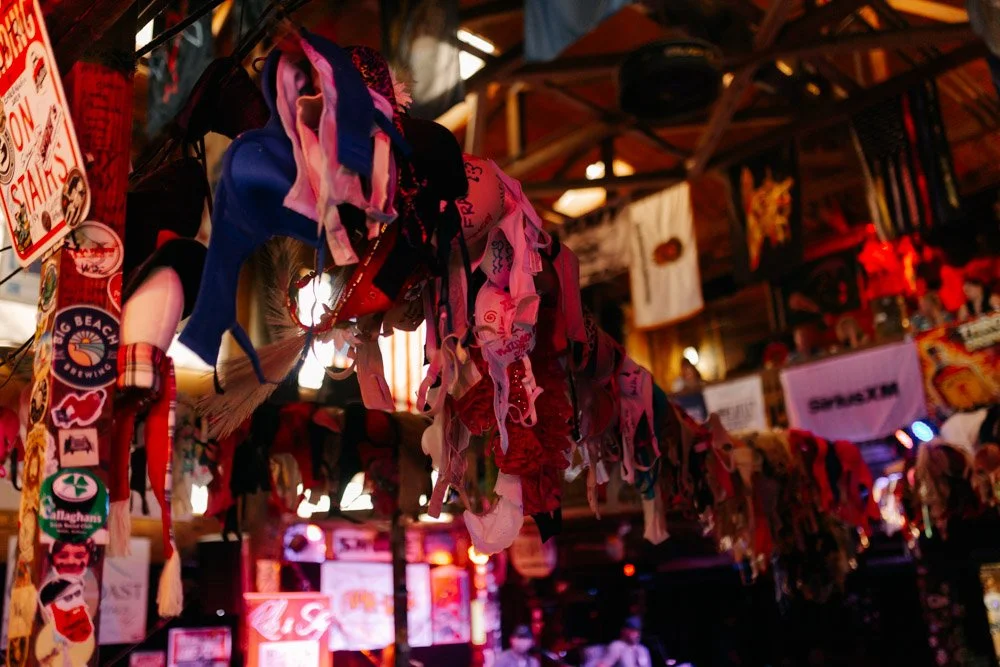 Clothing and hats hanging from a rack inside a bar or restaurant, decorated with various stickers and banners.