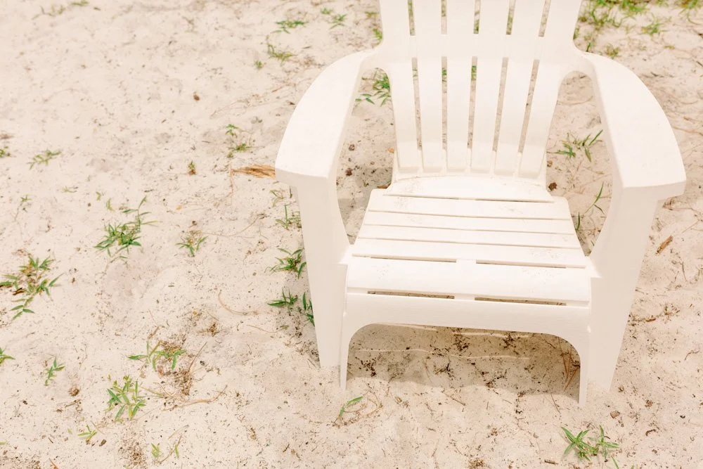 A white plastic beach chair on sandy ground with small patches of green grass.