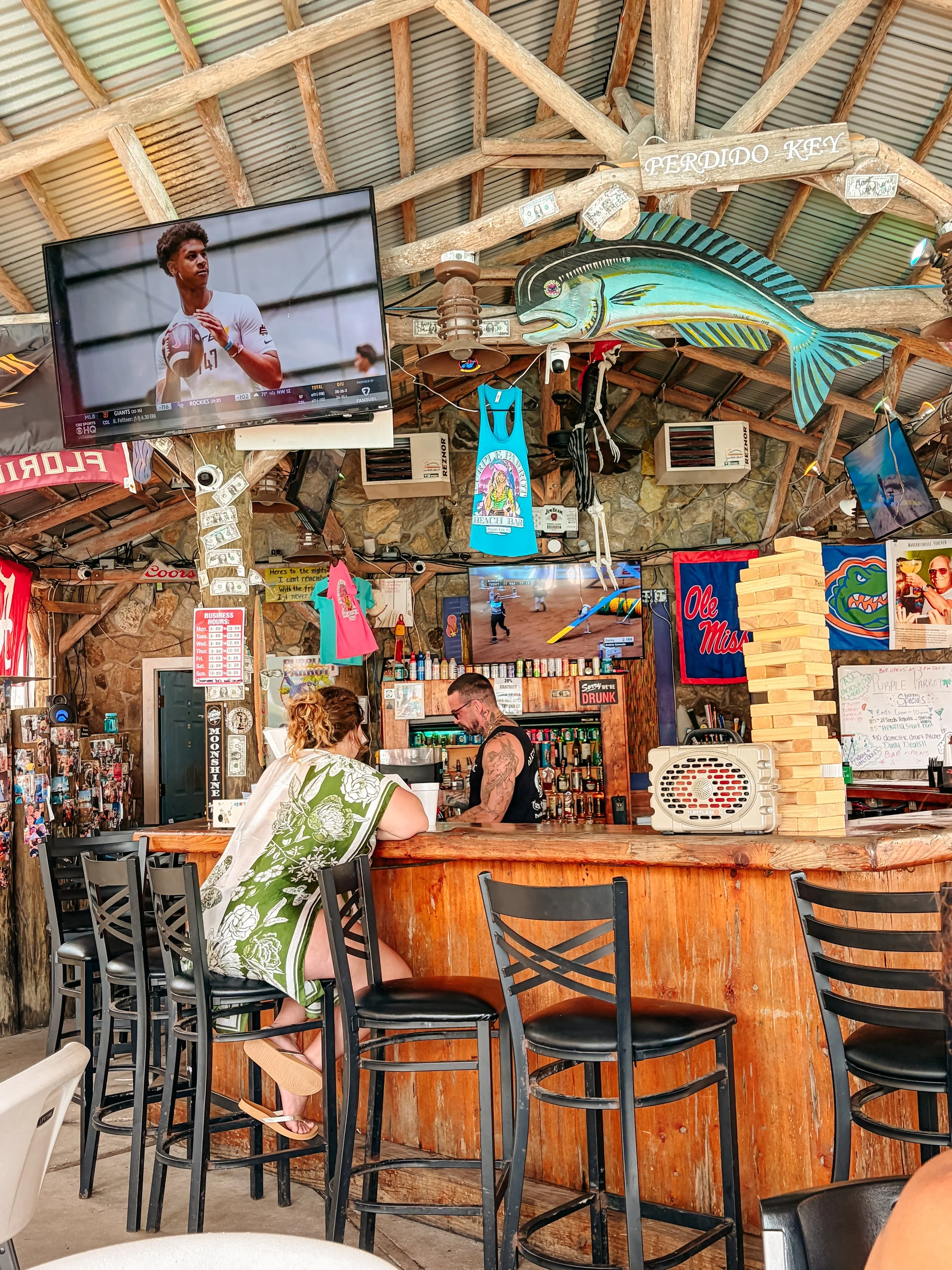 Bar area with high chairs and two people sitting. A television mounted on the wall shows a sports event. Decorations include a large fish sculpture, signs, flags, and hanging clothing in a rustic setting with wooden beams.