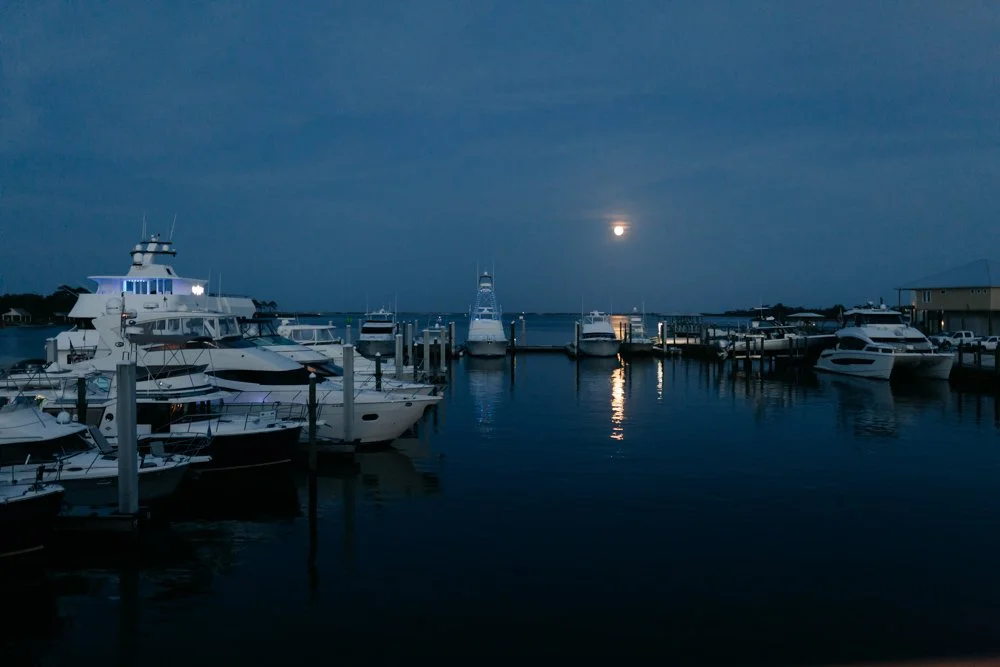 A marina at night with calm water, several yachts docked, moon partially covered by clouds, and a reflection on the water.