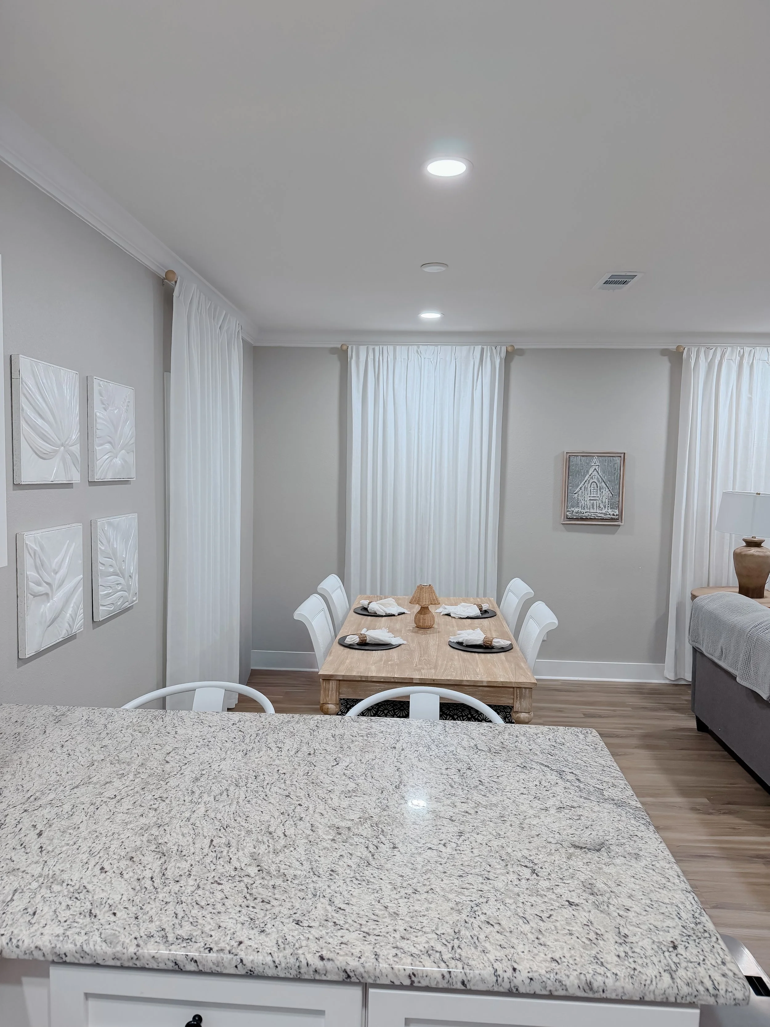 A kitchen and dining room view, showing a granite countertop in the foreground, a dining table with four chairs, and two windows with white curtains in the background.