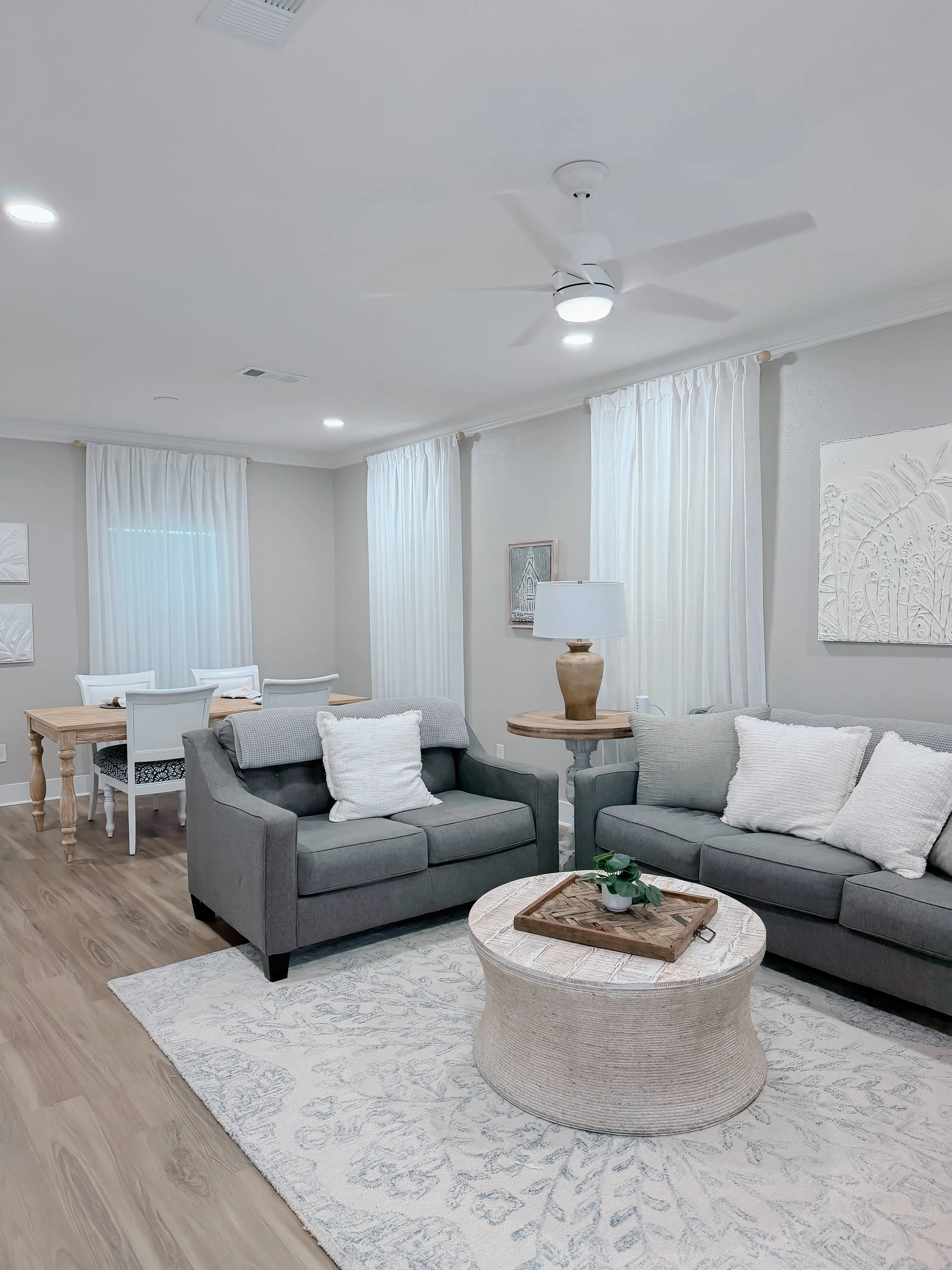Living room with gray sofas, white pillows, wooden coffee table, white curtains, artwork on wall, ceiling fan, and hardwood floors.