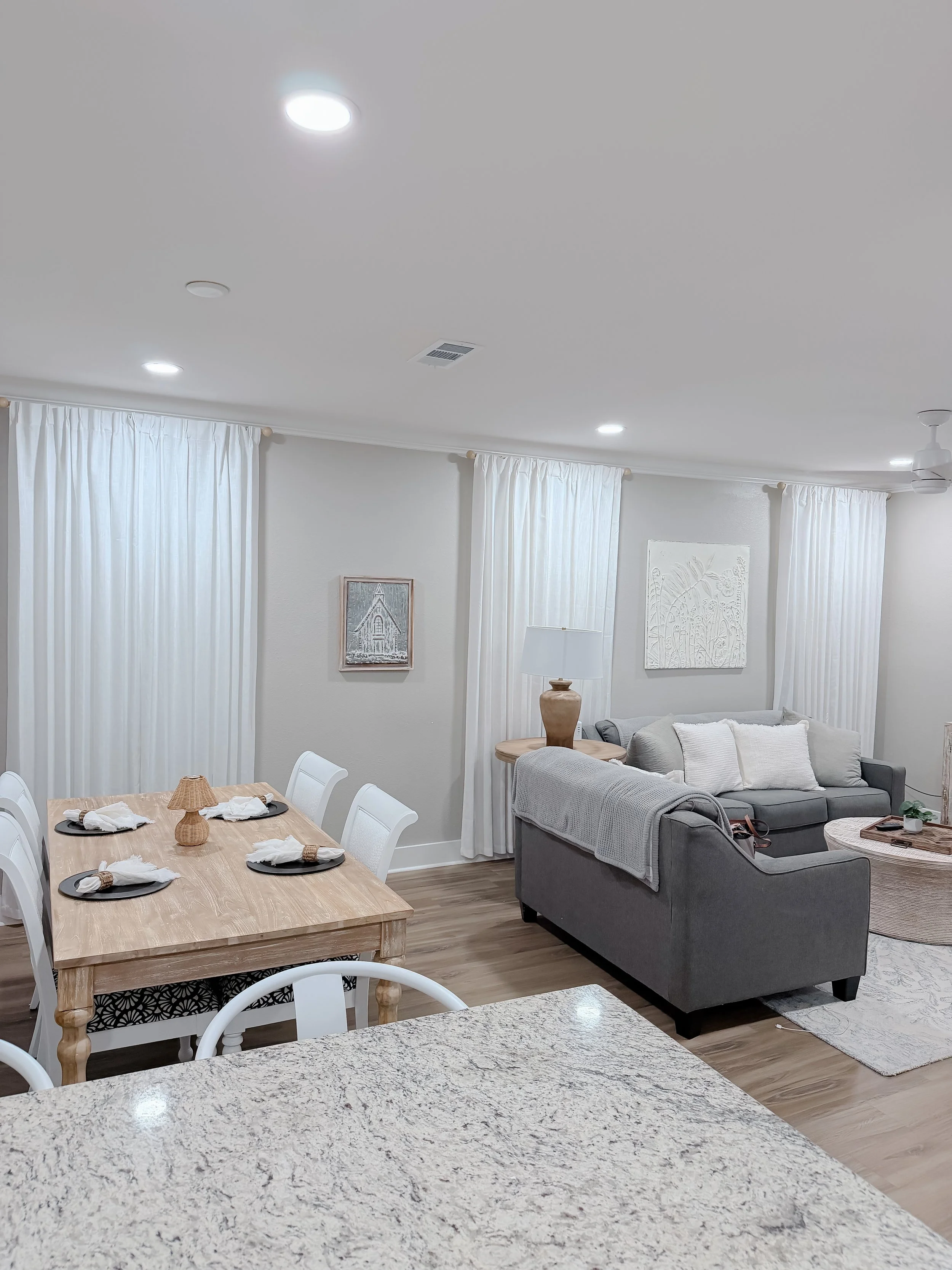 A modern, bright living room and dining area with white curtains, a gray sofa, a wooden dining table with white chairs, and decorative wall art.
