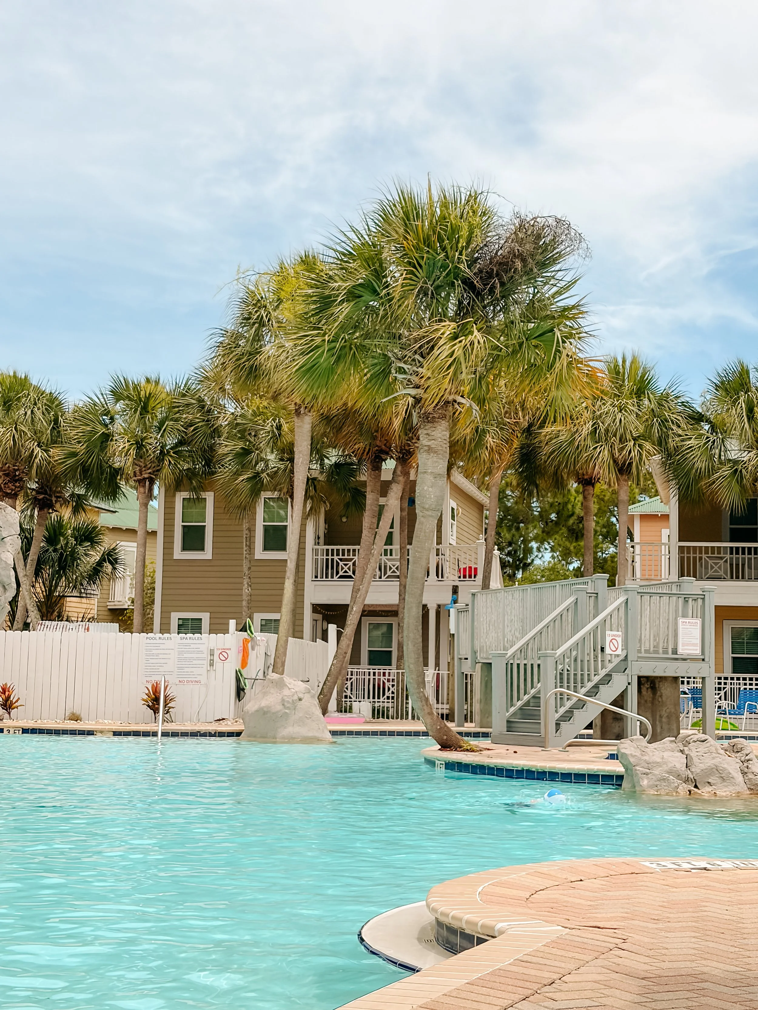 Swimming pool with light blue water, surrounded by a beige tiled deck, with palm trees and residential buildings in the background.