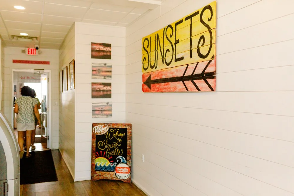 Interior of a restaurant or cafe with beach-themed decor, including a large sunset sign with an arrow and a Welcome to Sunset Grillboard.