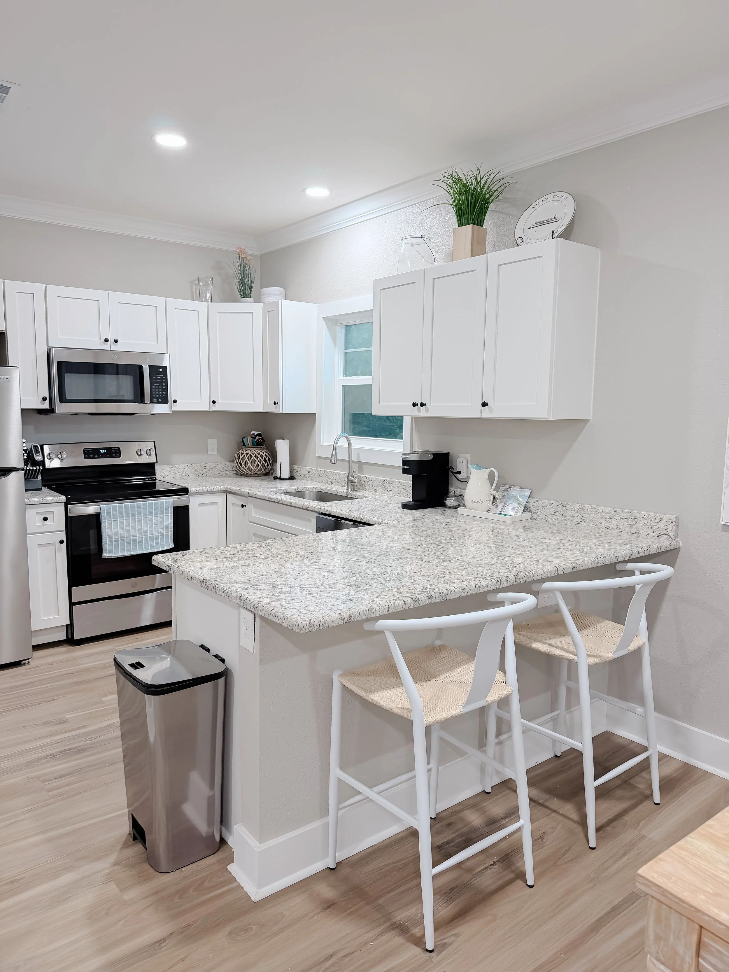 Modern kitchen with white cabinets, granite countertops, stainless steel appliances including a microwave and oven, a kitchen island with two white chairs, a trash can, small decorative items, and a window