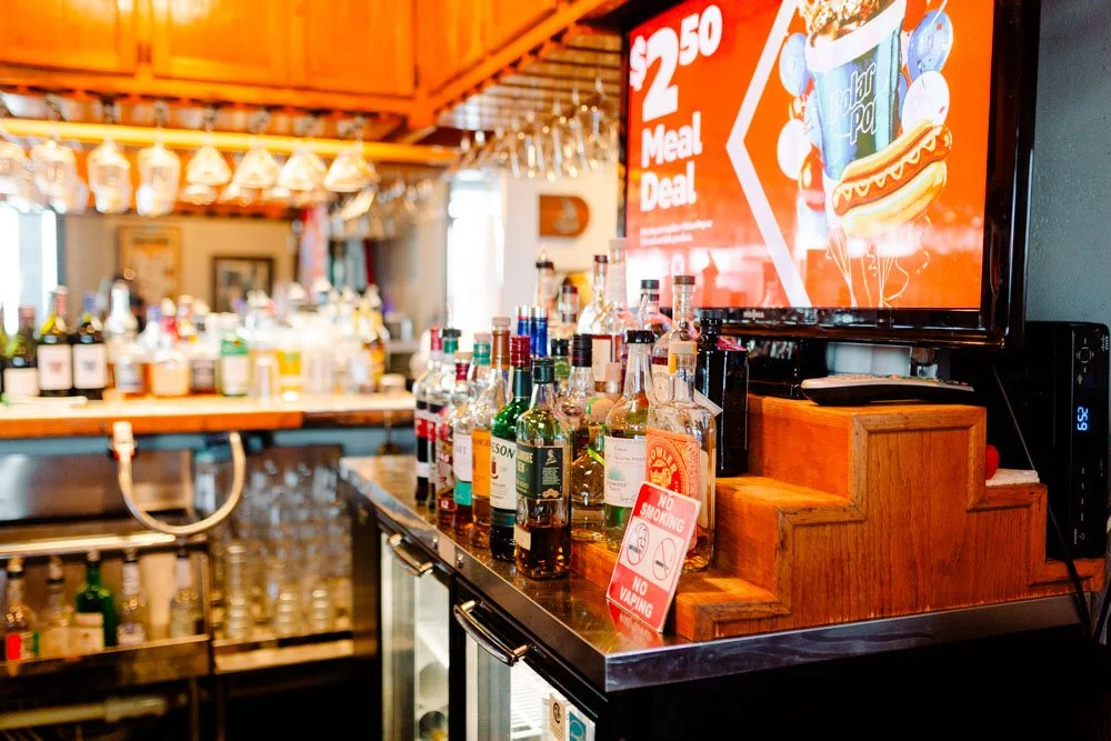 Bar counter with various liquor bottles, a digital menu screen displaying a meal deal, and a no smoking and no vaping sign.