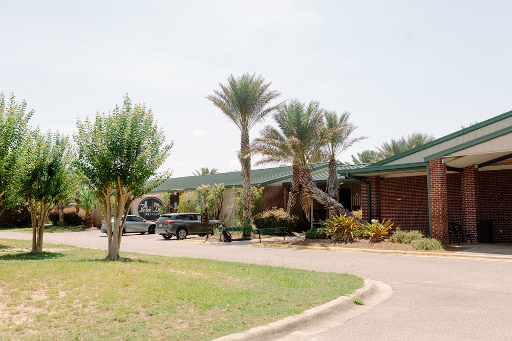 A single-story brick building with a green metal roof, surrounded by trees including palm trees, with a parking lot in front containing cars and a motorcycle, and a gravel walking path.