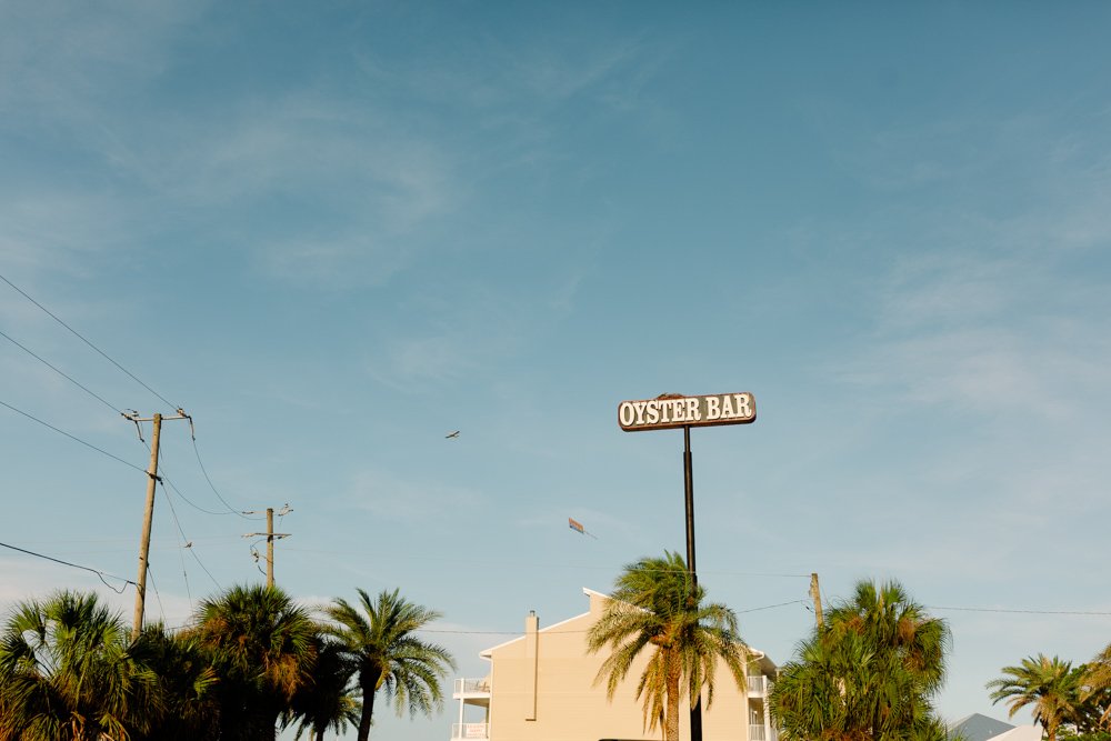 Sign that reads 'Oyster Bar' above a white building surrounded by palm trees under a blue sky.