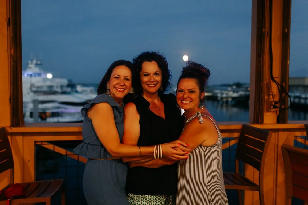 Three women smiling and hugging each other at a marina during the evening, with boats and a full moon in the background.