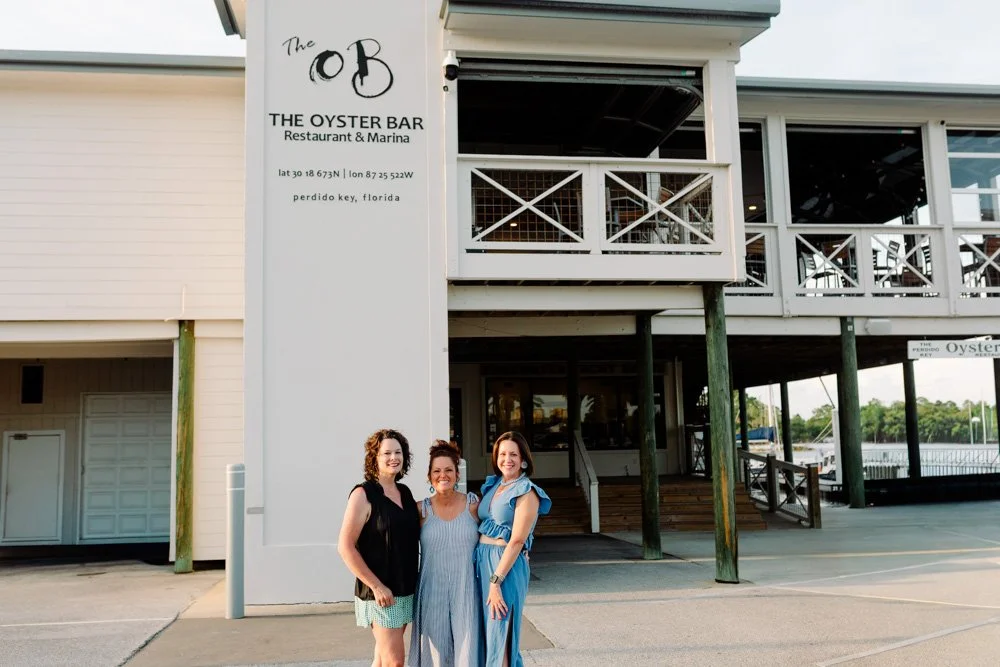 Three women standing in front of The Oyster Bar restaurant and marina in Perdido Key, Florida.