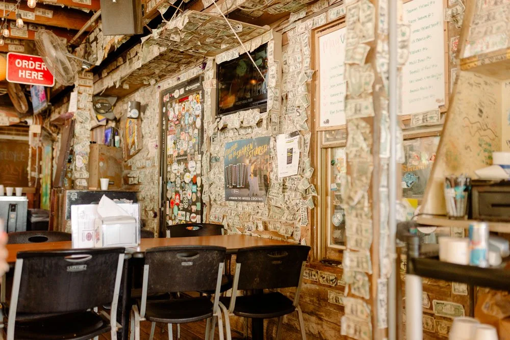 Inside a restaurant with dollar bills taped all over the walls and ceiling, wooden tables, black chairs, and various posters and signs on the wall.