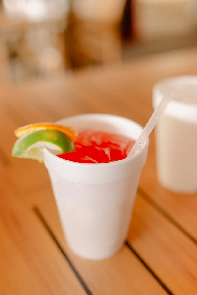 A white disposable cup filled with a red beverage, garnished with a slice of lime and lemon and a white straw. Another cup is partially visible in the background on a wooden table.