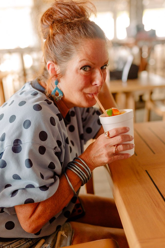 Woman with curly red hair in a bun, wearing a polka dot shirt and large earrings, drinking from a cup with a straw at a wooden table in a sunlit cafe.