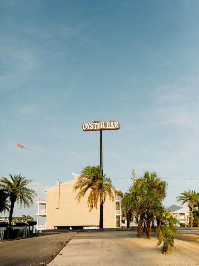A street view with palm trees and a building in the background, featuring a tall sign that reads 'OYSTER BAR' against a blue sky.