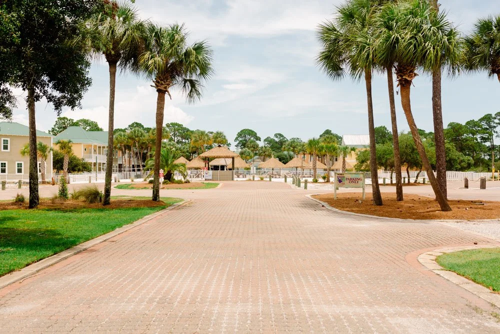 A paved road flanked by palm trees leading to a residential or resort area with buildings, green lawns, and a thatched roof structure under a partly cloudy sky.