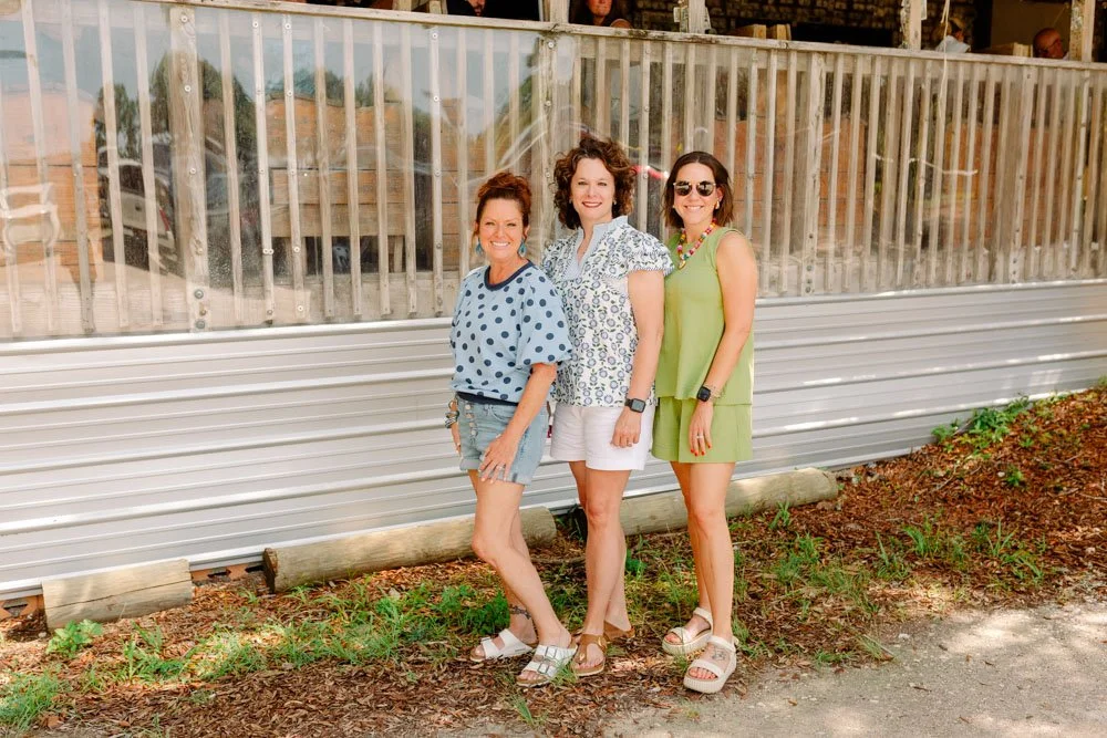 Three women standing outdoors in front of a metal and plastic fence, smiling, wearing casual summer clothes.