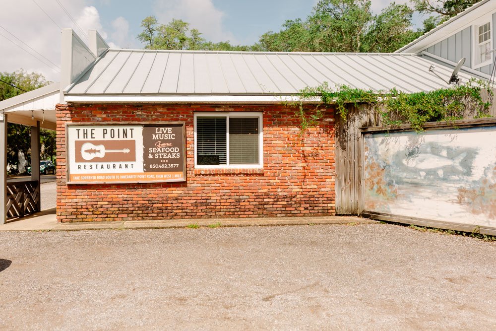 Brick building with a sign that reads "The Point Restaurant," marketing live music, seafood, and steaks with a phone number, and featuring a guitar graphic. There is a window with blinds and some greenery above the building.