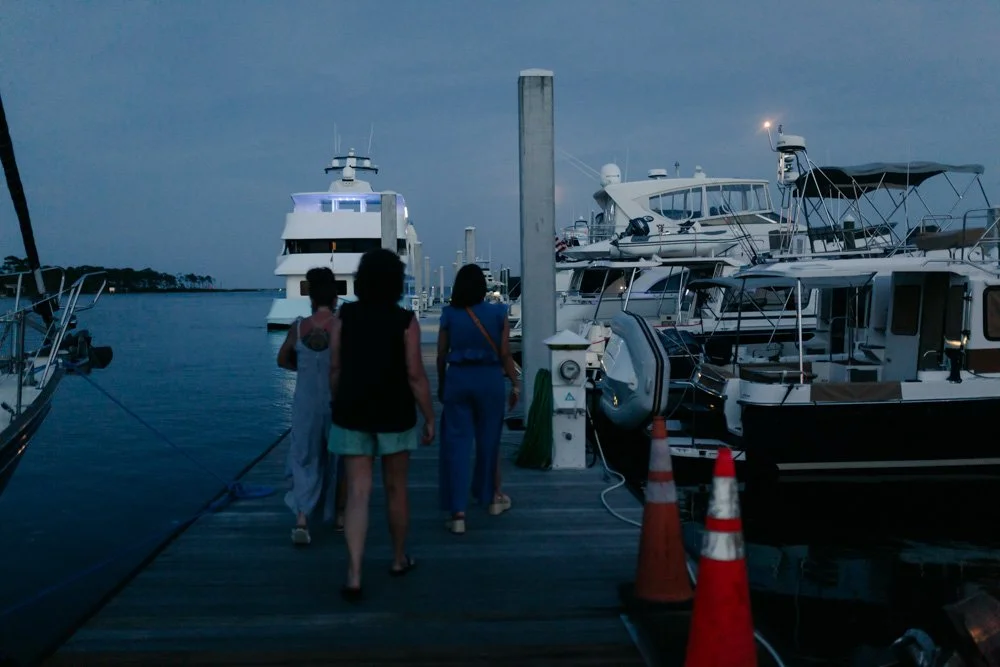 People walking on a dock with boats and yachts moored on either side, with a large yacht in the background, during twilight or early evening.