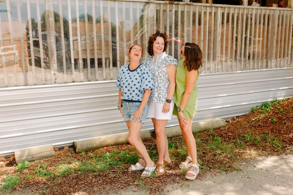 Three women standing and laughing outdoors in front of a metal and wooden fence.