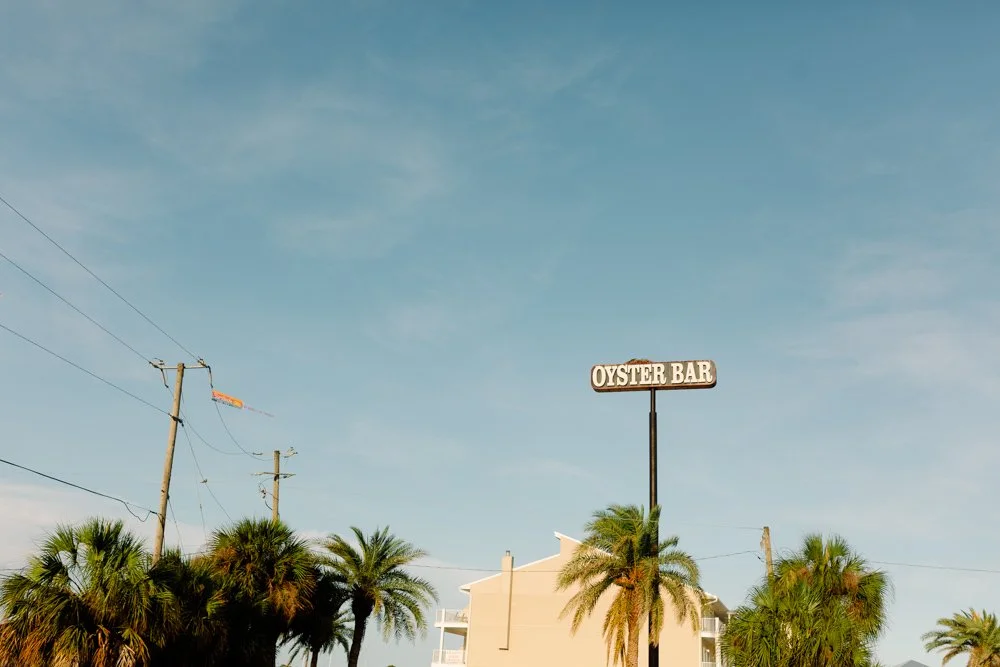 A clear sky above a sign that reads 'Oyster Bar,' with palm trees and a building in the background.