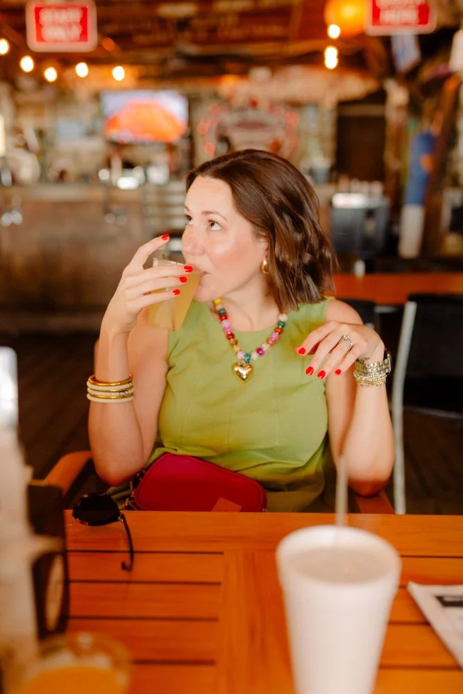 A woman in a green top and colorful jewelry drinks a beverage with a straw at a cozy, rustic bar or restaurant. She has short brown hair and red nail polish, and there are drinks and sunglasses on the table in front of her.