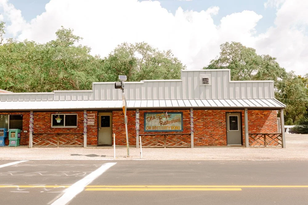A small brick building with a silver metal roof, two doors, a window, and a painted sign. There are two recycling bins on the left, with a gravel parking lot and trees in the background.