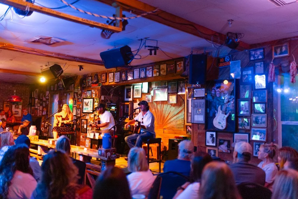 A live music performance in a cozy bar with string lights, framed photos on the wall, and an audience seated at tables.
