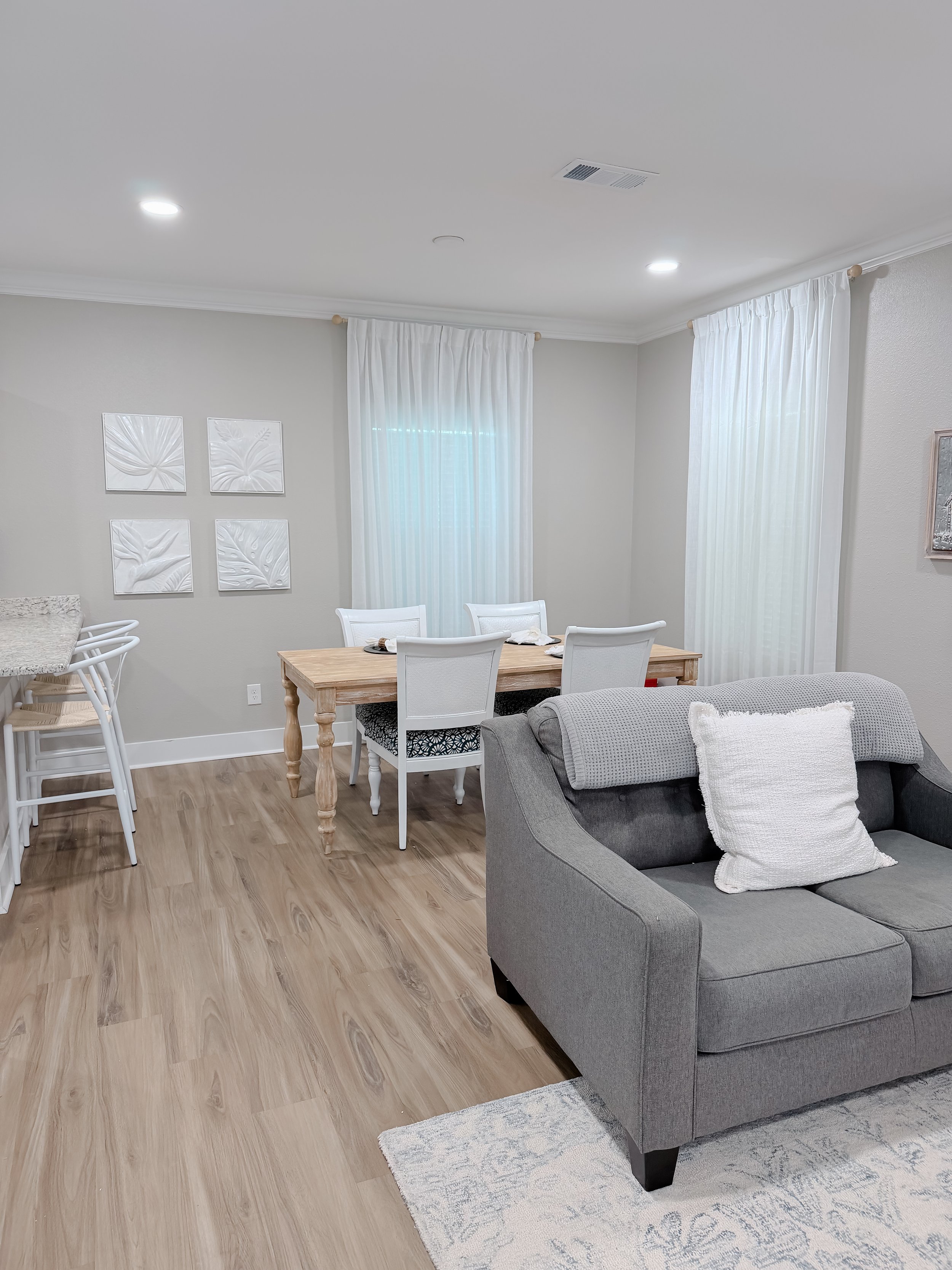 A living room with a gray sofa, a wooden dining table with white chairs, and a white side bar. White curtains cover the windows, and four white textured wall art pieces are on the back wall. The room features light wood flooring, a small rug, and cei