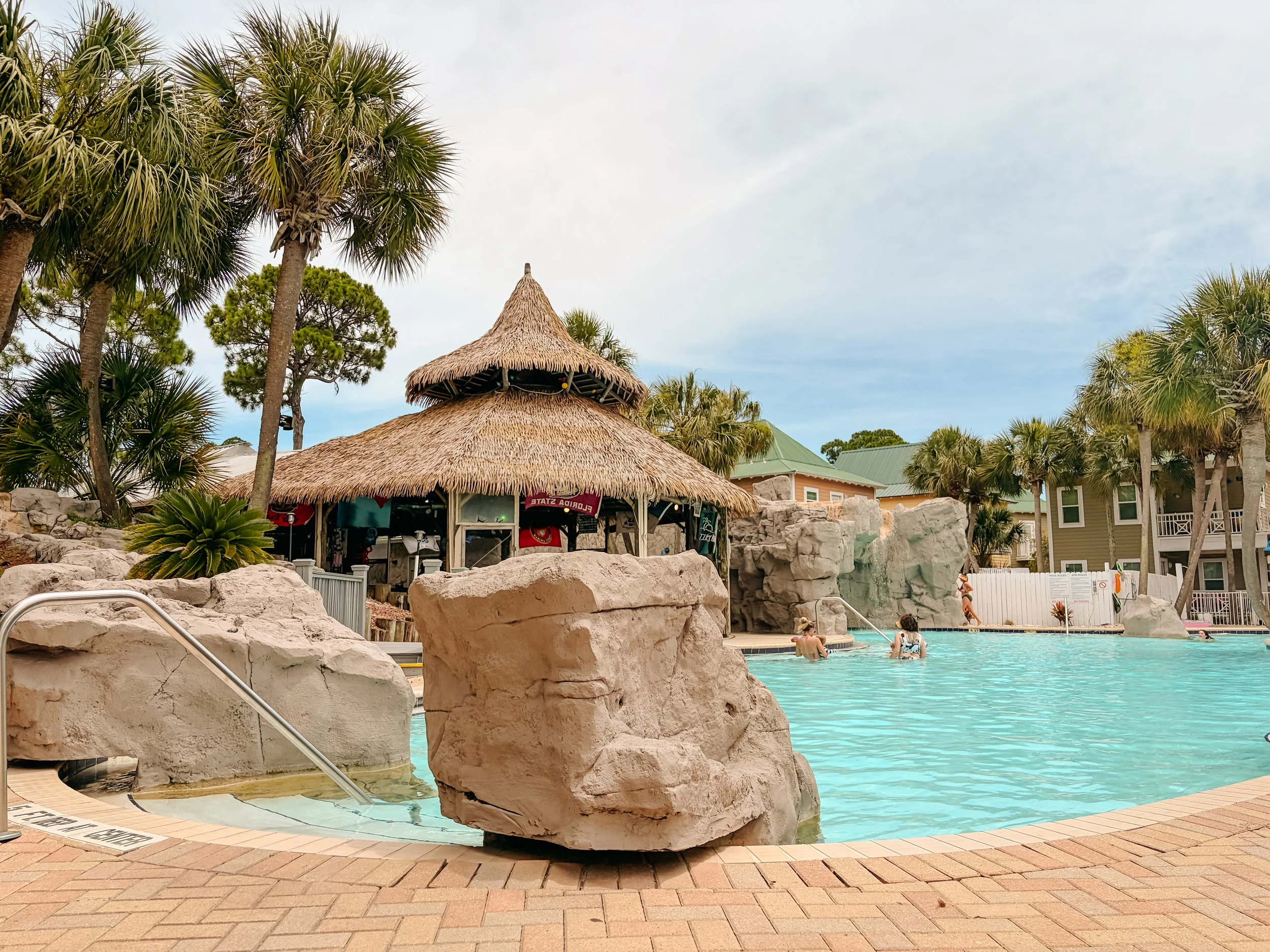 A swimming pool area with a thatched-roof tiki hut, surrounded by rocks and palm trees. Several people are in the pool, and there are buildings with green roofs in the background.