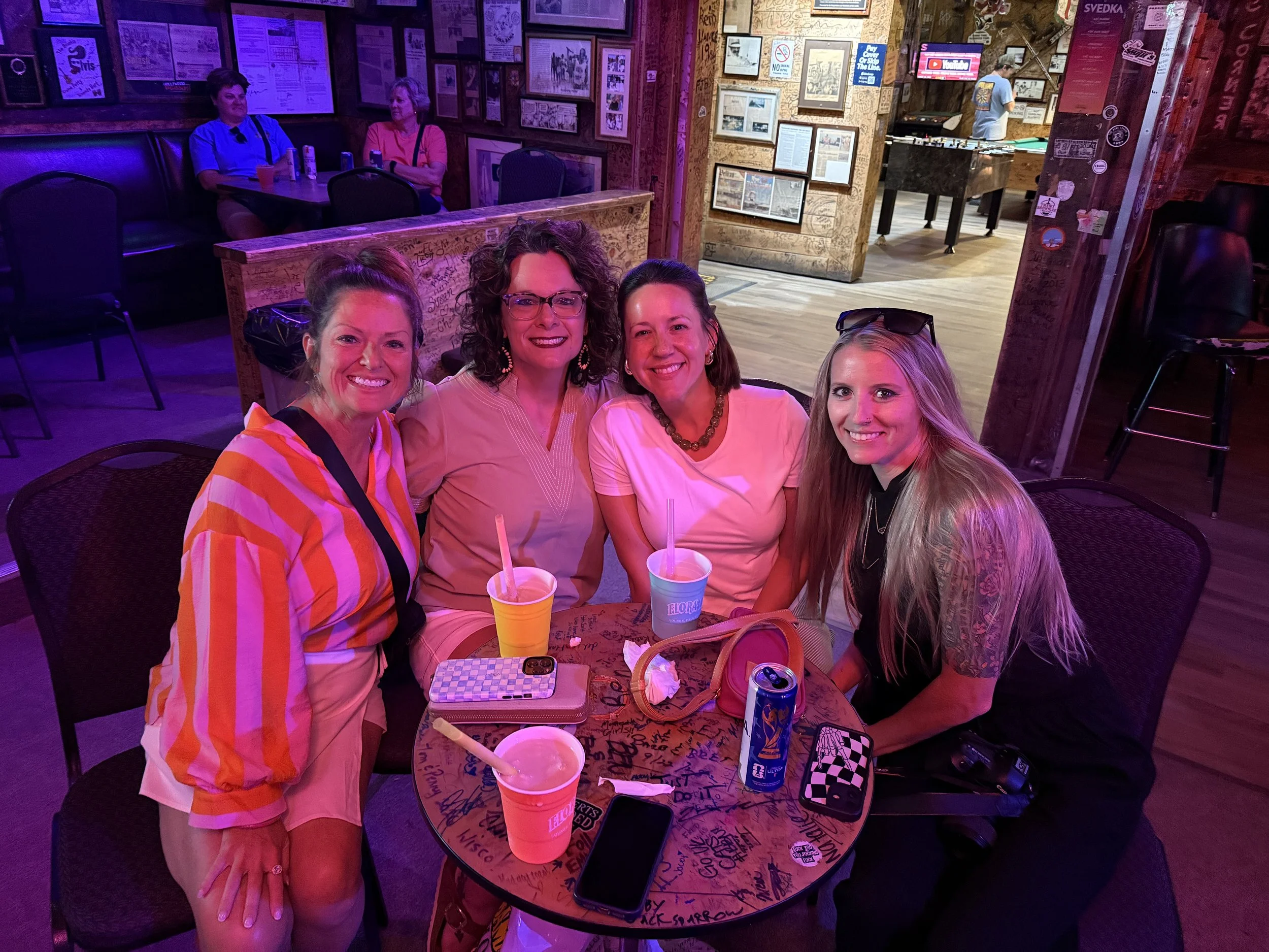 Four women sitting around a table in a bar or restaurant, smiling at the camera. The table has drinks, a phone, and various items on it. The background has decorated walls with framed pictures and signs, and a foosball table can be seen.