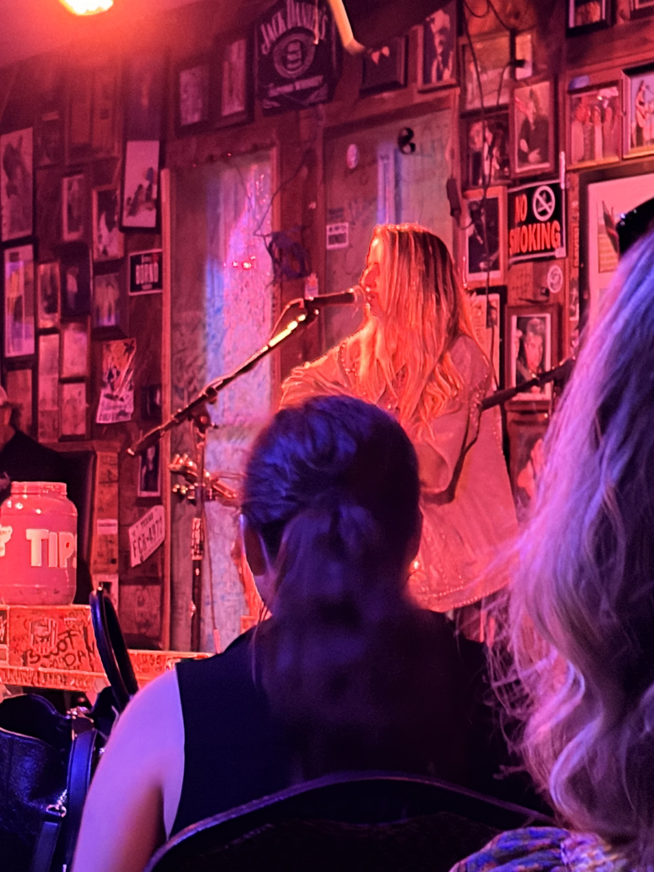 Person singing on stage in a dimly lit bar with red and purple lighting, surrounded by framed photos and posters on the wall, with a 'No Smoking' sign visible.