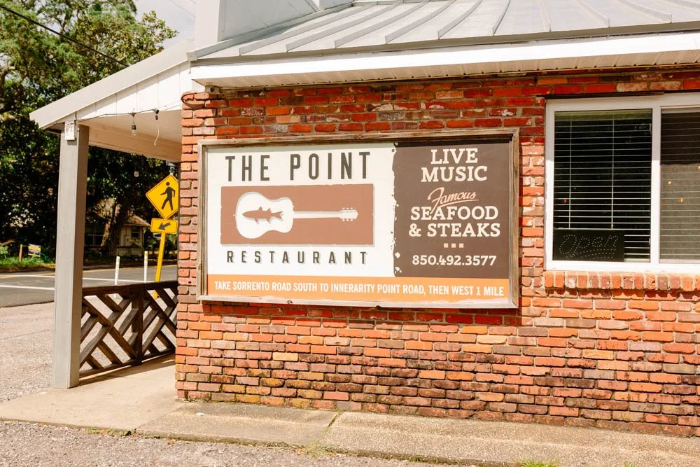Sign on brick building advertising The Point restaurant with live music, seafood, and steaks, including phone number 850.492.3577 and driving directions.