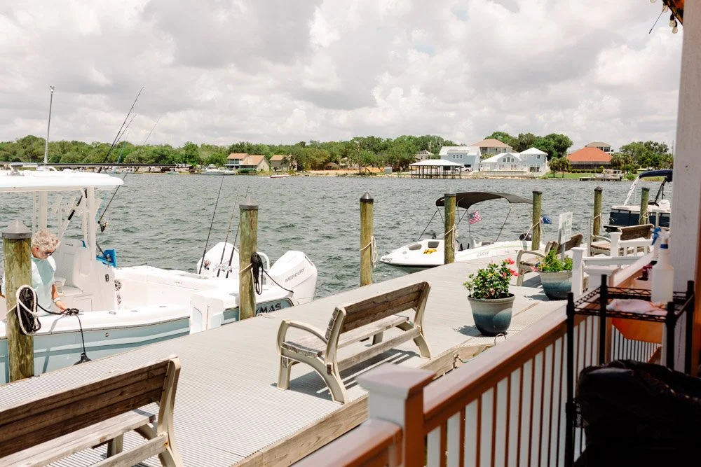 View of a waterfront dock with boats, a bench, potted plants, and houses on the opposite shore under a cloudy sky.