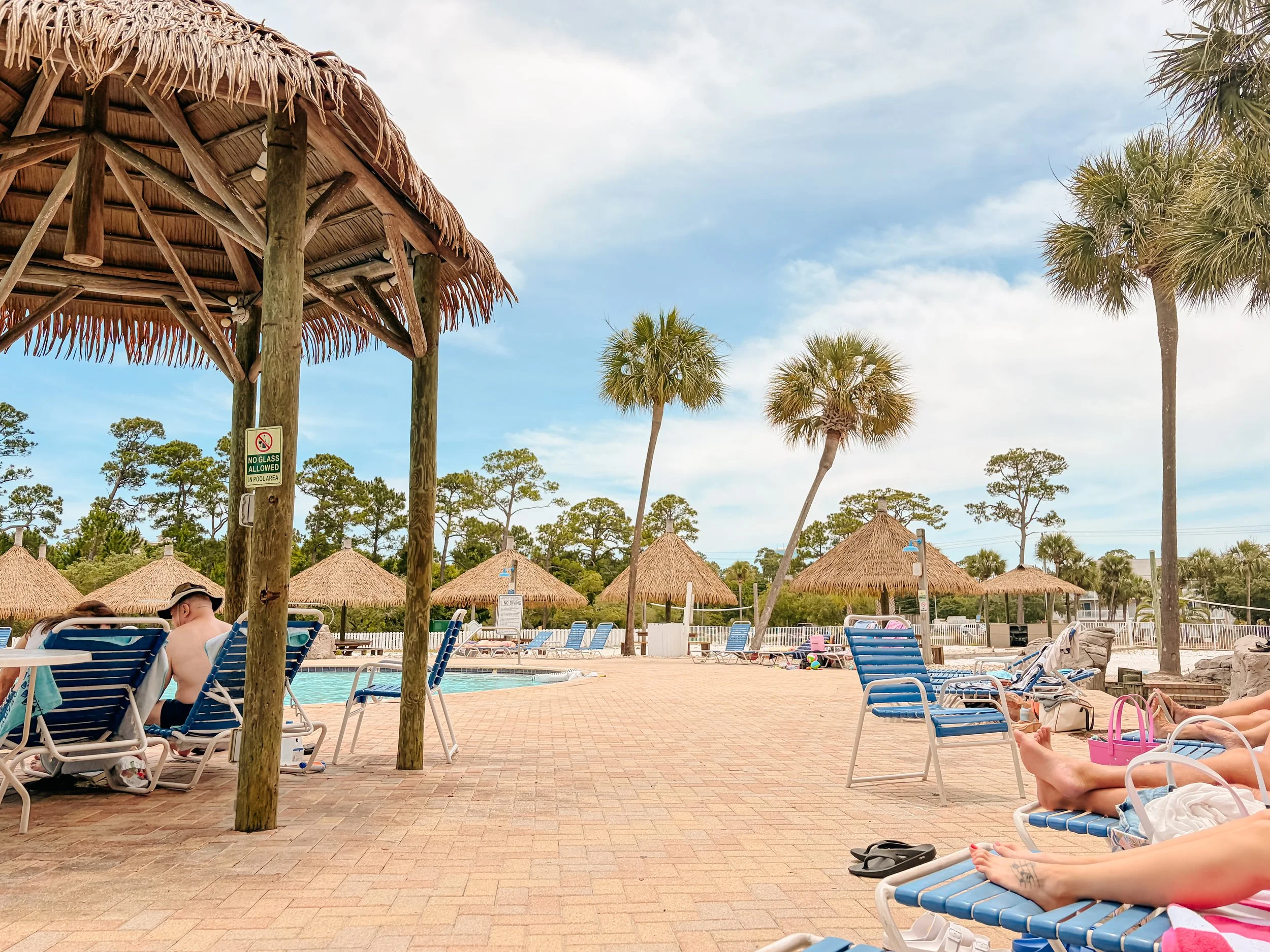 People relaxing on lounge chairs and enjoying a swimming pool at a tropical resort with thatched roofs, palm trees, and a partly cloudy sky.