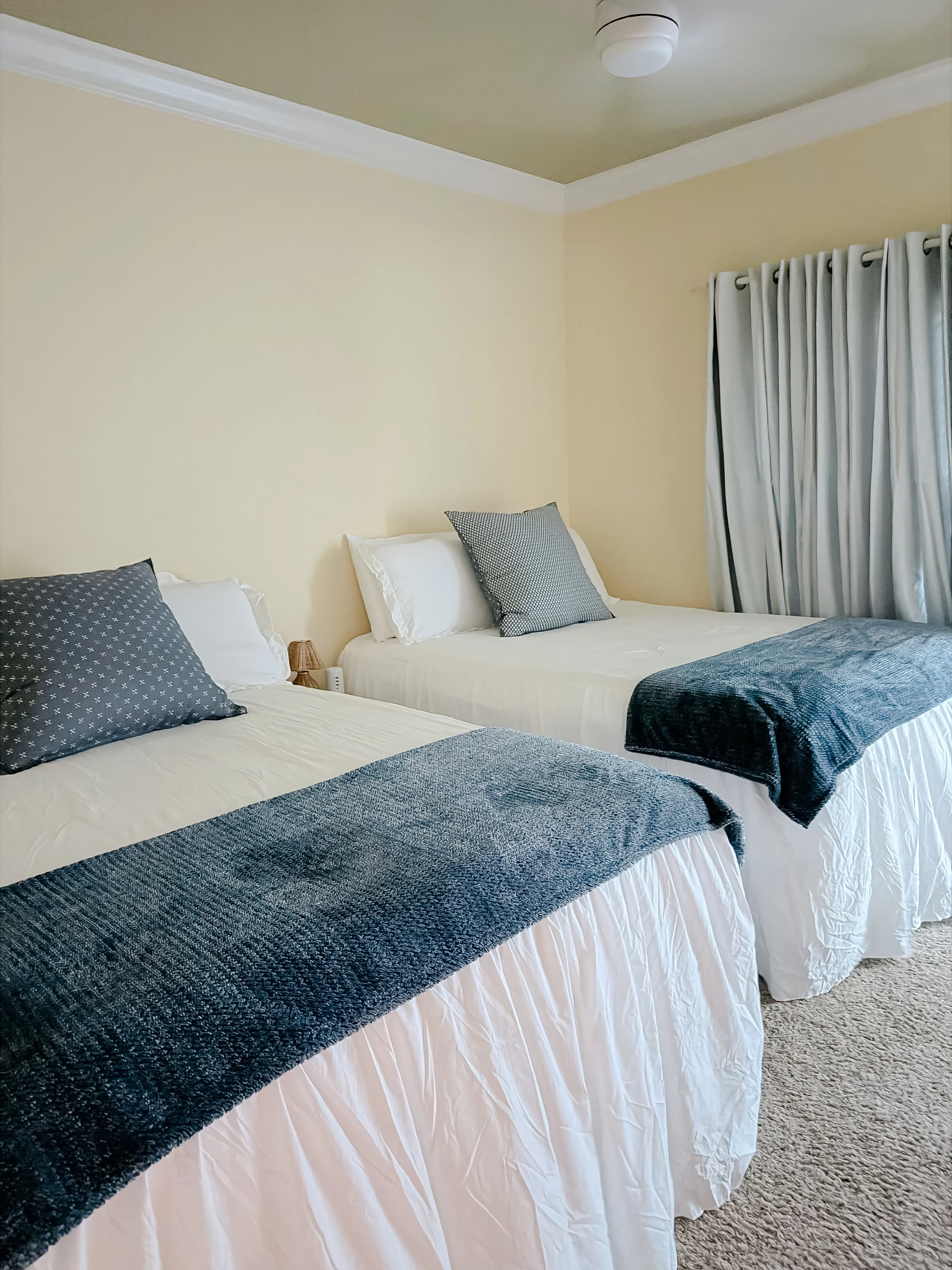 Two neatly made beds with white sheets, each topped with dark gray and light gray throw blankets and pillows, in a bedroom with beige walls, white trim, white curtains, and a carpeted floor.