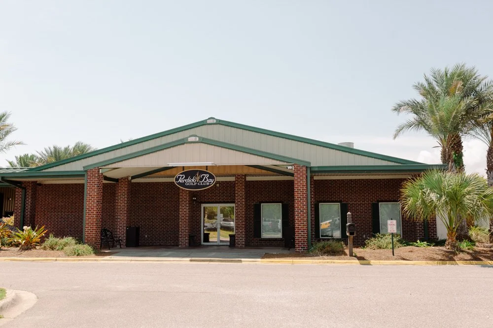 Exterior view of Perdido Bay Golf Club with brick building, green roof, palm trees, and parking lot in Florida.