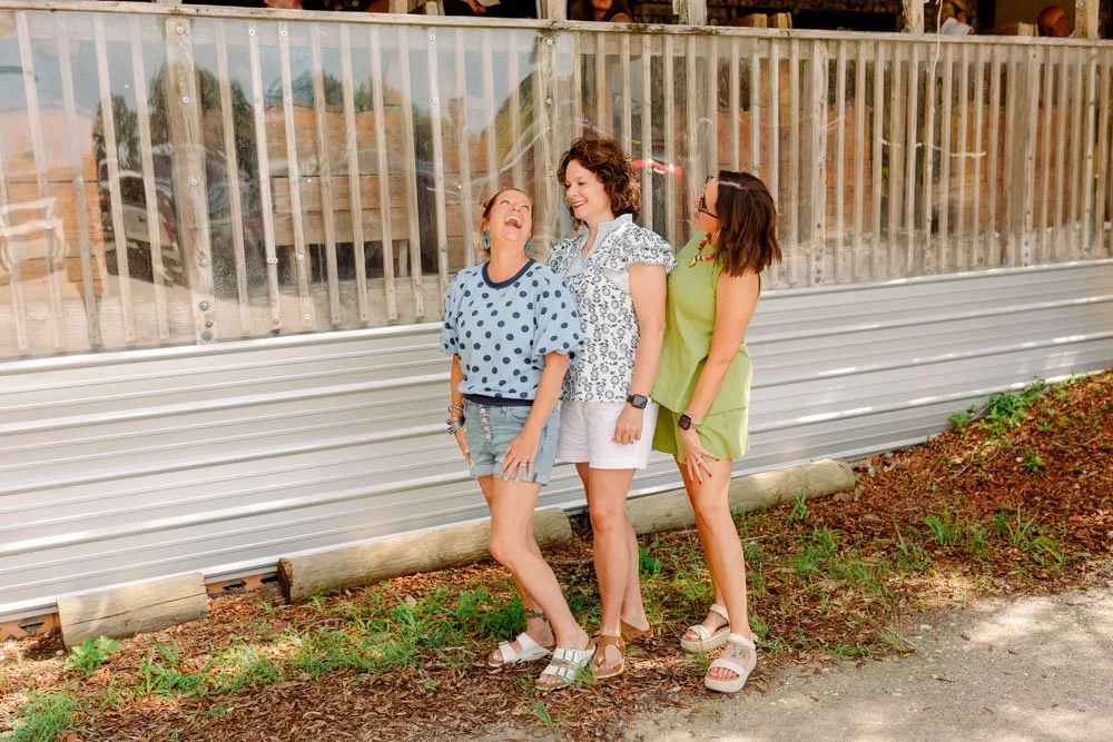 Three women stand outside near a wooden fence, laughing and smiling together.