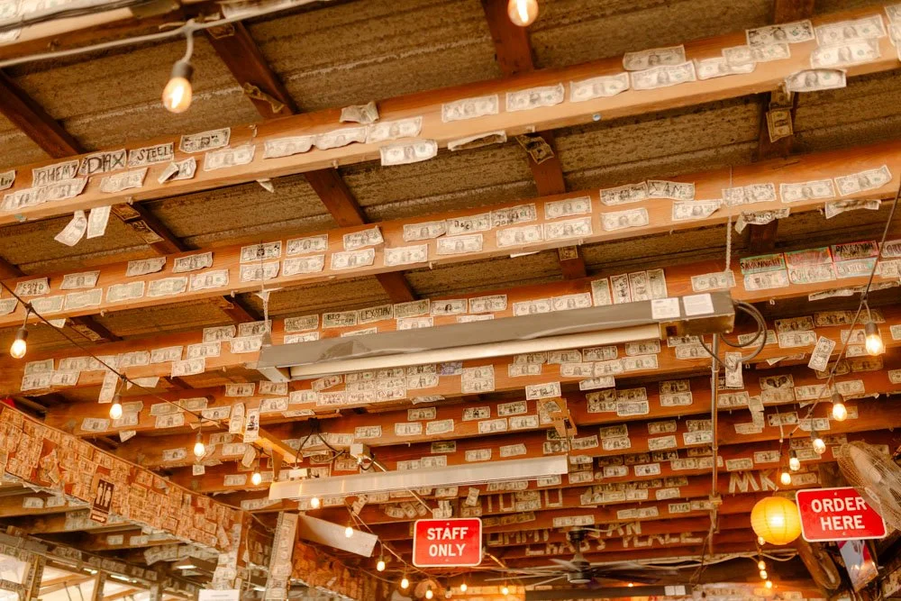Wooden ceiling with dollar bills attached to the beams and hanging string lights; red signs indicating 'STAFF ONLY' and 'ORDER HERE' in a restaurant or cafe.