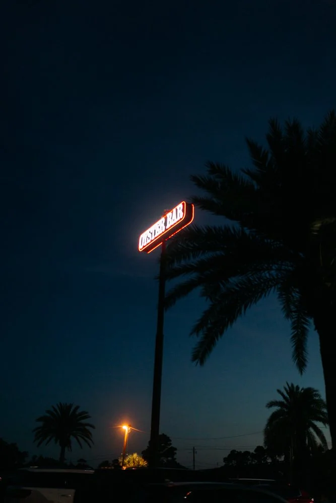 Nighttime scene with a tall sign reading 'OASIS BAR & BQ' illuminated in red, palm trees, and a sky with a faint glow near the horizon.