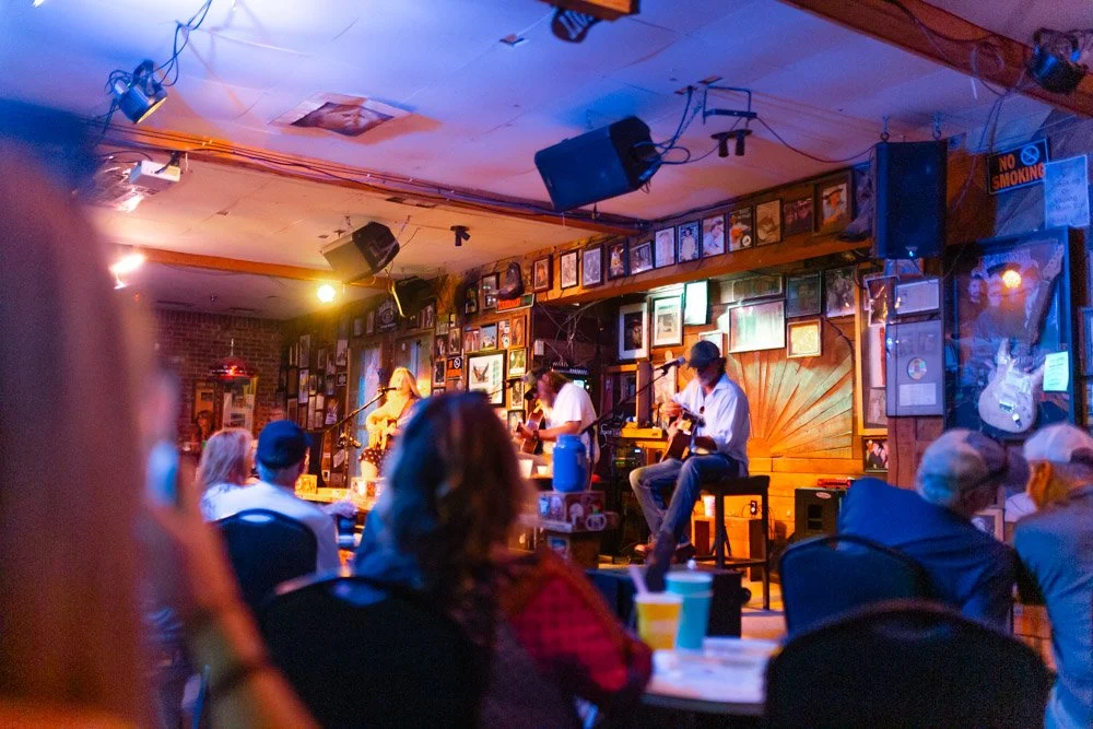 Live music performance at a bar with a small stage, featuring two guitarists and a singer, decorated with framed photos on the wall, surrounded by patrons sitting and enjoying the show.