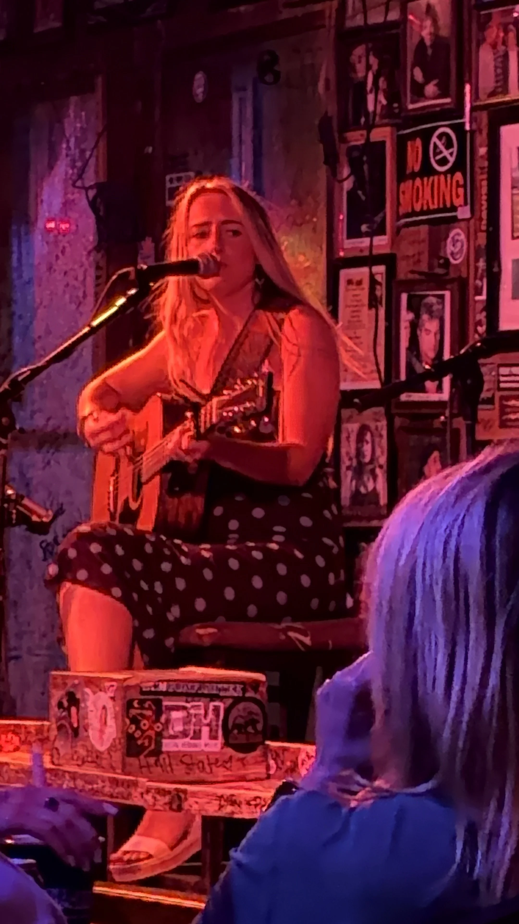 A woman with long blonde hair singing and playing an acoustic guitar on stage, wearing a striped top and polka dot pants, with mural-style graffiti and framed portraits on the wall behind her, and an audience watching in front.