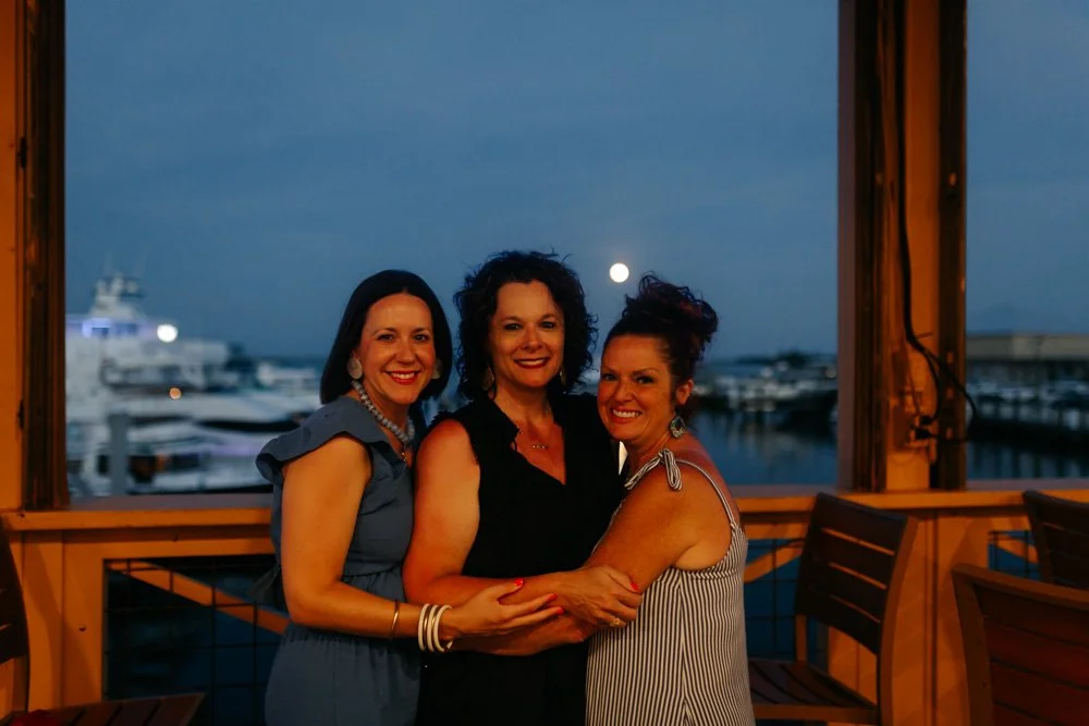 Three women smiling and hugging each other on a balcony overlooking a marina with boats at night, with a Full Moon in the sky.