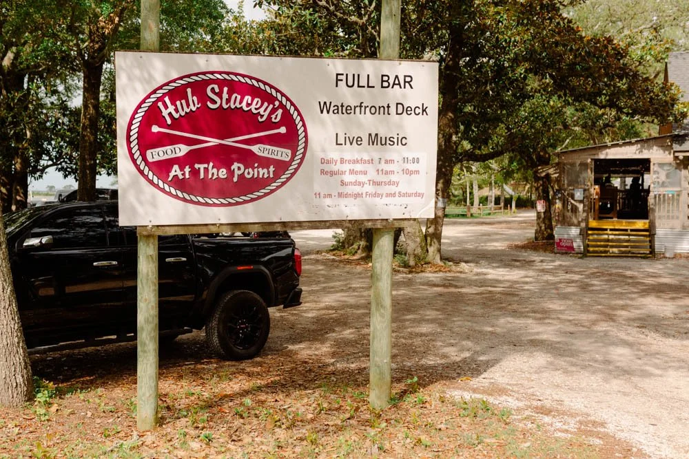 A signboard advertising Hub Stacey's At The Point, a waterfront bar with live music, full bar, and daily breakfast. The sign displays hours of operation and is mounted on two wooden posts in a parking area with trees and a small building in the background.
