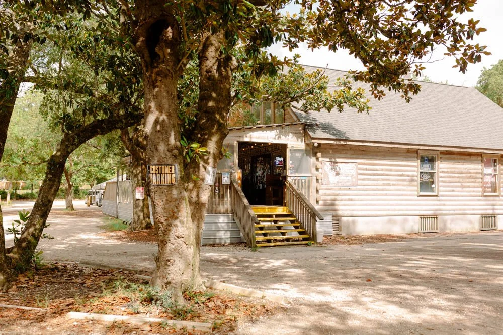 A rustic log cabin-style building with wooden stairs leading to the entrance, surrounded by trees with green leaves, on a gravel lot with a 'Customer Parking' sign on a tree.