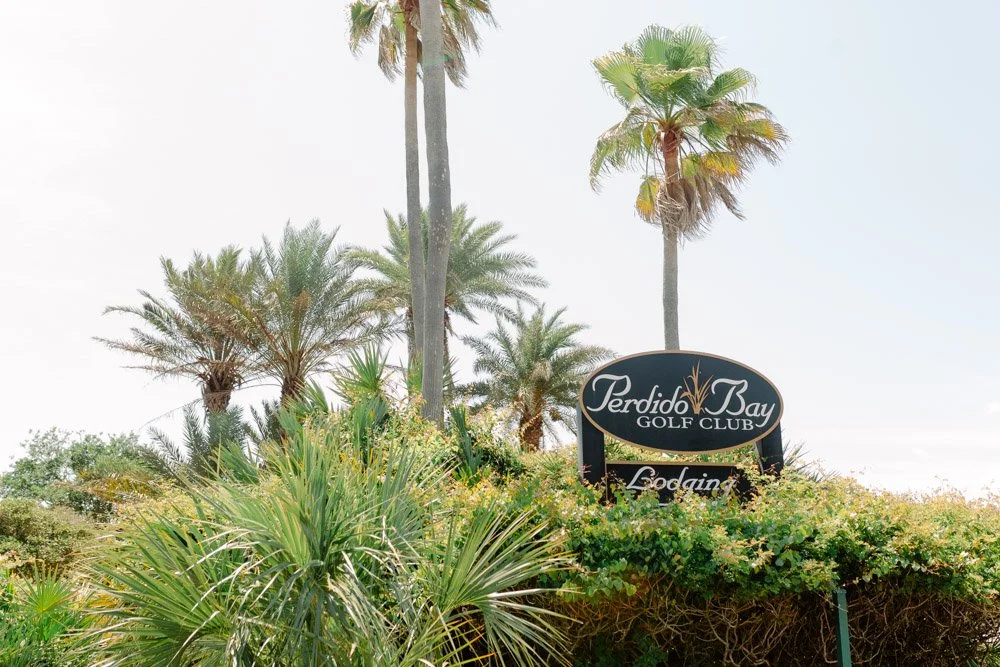 Sign for Perdido Bay Golf Club surrounded by palm trees and lush green plants in a tropical landscape.